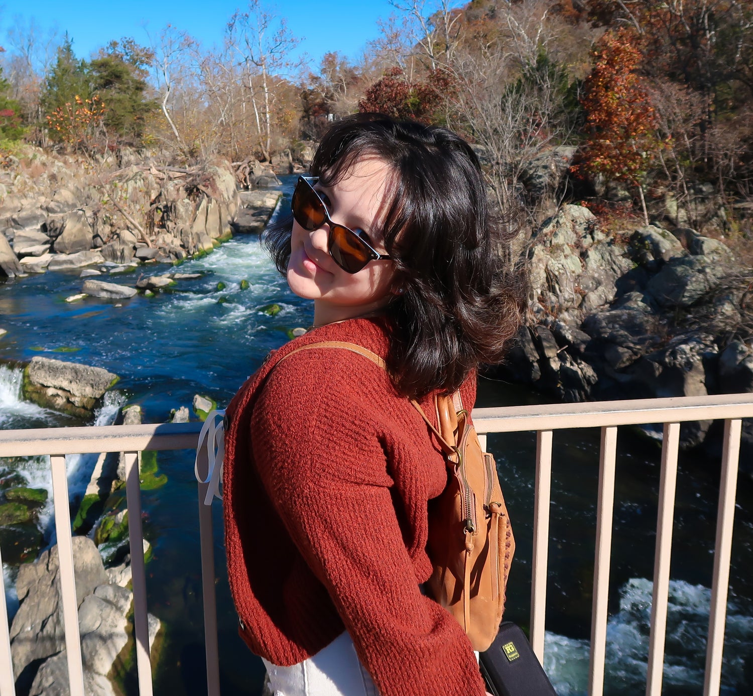 Person wearing sunglasses and a red sweater standing by a river with rocky landscape